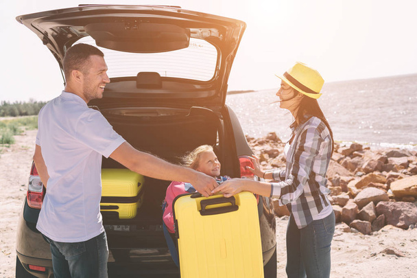 Young couple standing near the opened car boot with suitcases and bags. Dad, mom and daughter are traveling by the sea or the ocean or the river. Summer ride by automobile. - Фото, изображение