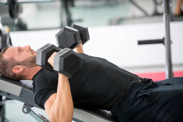 Young handsome adult male doing dumbbell rows on bench in modern gym - Foto, afbeelding