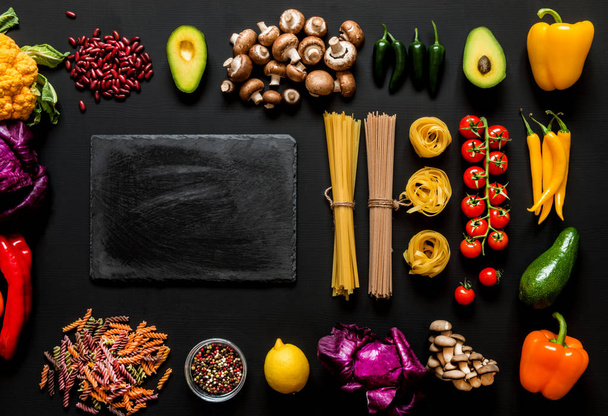 Different colorful Fresh raw ingredients for healthy vegetarian cooking on a black background with blackboard and free copy space. Flat lay, top view. - Фото, зображення