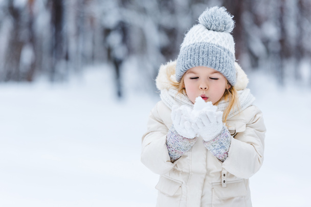 portrait of adorable kid blowing onto snow ball in hands in winter park - Photo, Image