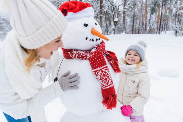 happy mother and daughter playing with snowman in winter park - Photo, Image