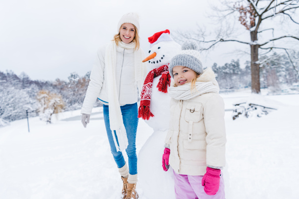 happy mother and daughter smiling at camera while standing with snowman in winter park - Photo, Image