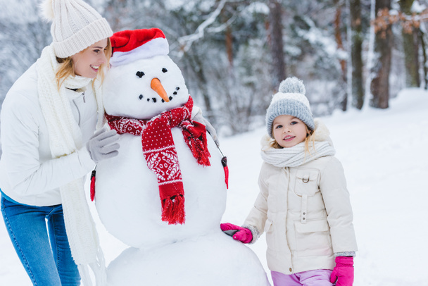 красива щаслива мати і дочка, що стоїть зі сніговиком в зимовому парку
 - Фото, зображення