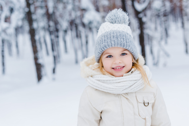 portrait of cute little child in hat and scarf smiling at camera in winter park - Valokuva, kuva