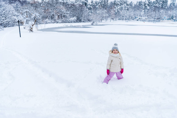 adorável criança feliz de pé na neve e sorrindo para a câmera no parque de inverno
   - Foto, Imagem