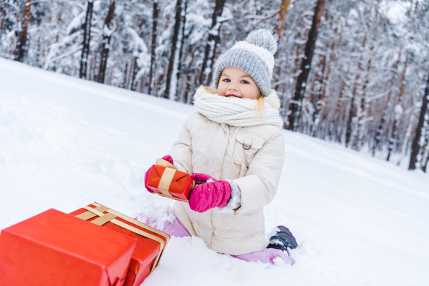 bambino felice in abiti invernali seduto sulla neve con scatole regalo e sorridente alla macchina fotografica
 - Foto, immagini