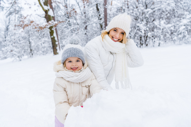 beautiful happy mother and daughter smiling at camera while making snowman in winter park - Fotoğraf, Görsel