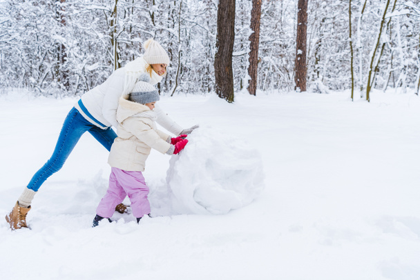 side view of happy mother and daughter making snowman in winter park - Foto, Imagen