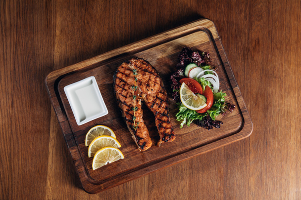 top view of delicious grilled salmon steak served on wooden board with lemon slices and salad - Foto, Bild