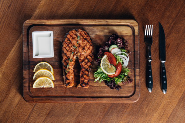 bovenaanzicht van gegrilde zalm steak, geserveerd op een houten bord met plakjes citroen en salade - Foto, afbeelding