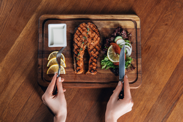 cropped shot of woman eating grilled salmon steak served on wooden board with lemon and lettuce - Photo, Image