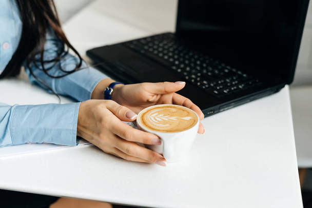 una chica en una camisa azul bebe un café fragante en un café, cerca de un ordenador portátil para el trabajo
 - Foto, Imagen