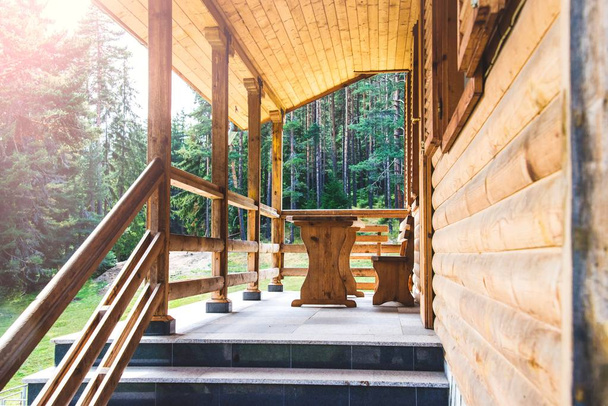 Oldalnézetből egy nyitott veranda előtt egy fából készült erdei házikó. Fenyves erdő alatt a nap sugarai a háttérben. - Fotó, kép