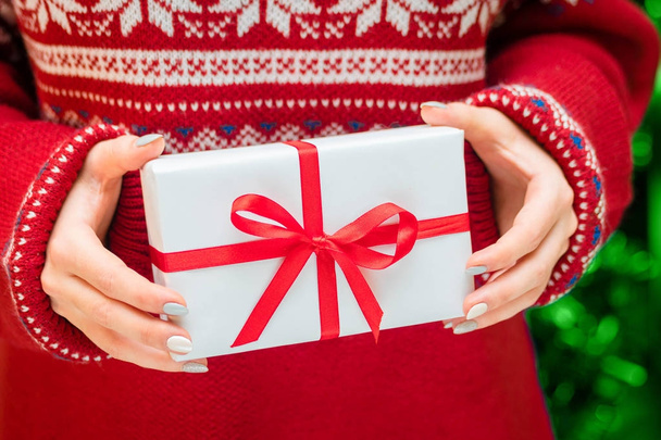 Woman showing beautiful white gift box with red ribbon- closeup of present on holiday sweater background and Christmas tree. - Фото, зображення