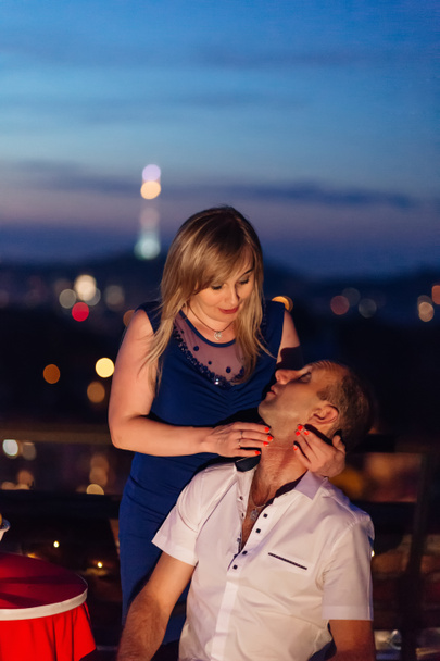 Женщина в великолепном платье обнимает своего мужа, который сидит на стуле на фоне вечернего города
. - Фото, изображение