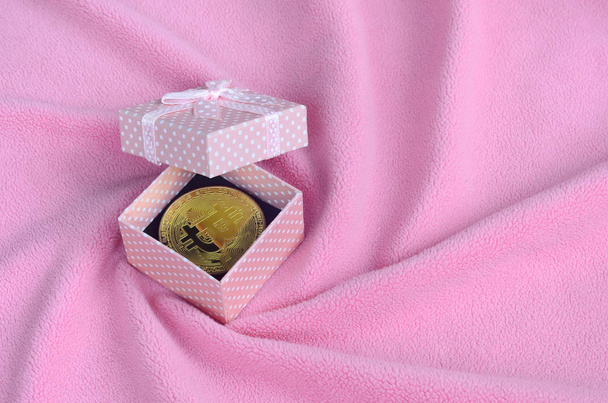 Золотой биткойн лежит в маленькой розовой подарочной коробочке с небольшим бантиком на одеяле из мягкой и пушистой светло-розовой шерстяной ткани с большим количеством рельефных складок
 - Фото, изображение
