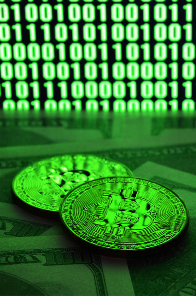 Two bitcoins lies on a pile of dollar bills on the background of a monitor depicting a binary code of bright green zeros and one units on a black background. Low key lighting - Фото, изображение