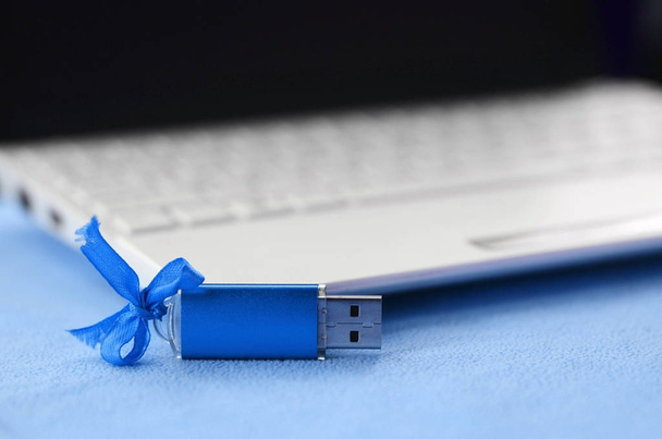 Λαμπρό μπλε usb κάρτα μνήμης flash με μπλε φιόγκο βρίσκεται σε ένα κάλυμμα από ύφασμα μαλακό και γούνινο φως μπλε φλις δίπλα σε ένα λευκό laptop. Κλασικά θηλυκές δώρων σχεδιασμός για μια κάρτα μνήμης - Φωτογραφία, εικόνα