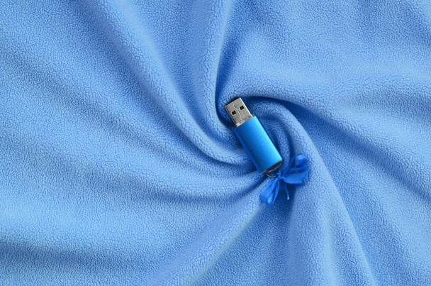 brillant blaue USB-Flash-Speicherkarte mit blauer Schleife liegt auf einer Decke aus weichem und pelzigem hellblauem Fleece-Stoff mit vielen Relief-Falten. Speichergerät im Frauendesign - Foto, Bild