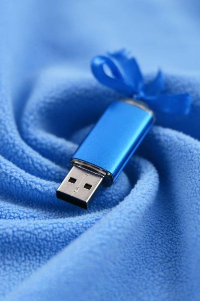 Λαμπρό μπλε usb κάρτα μνήμης flash με μπλε φιόγκο βρίσκεται σε ένα κάλυμμα από ύφασμα μαλακό και γούνινο φως μπλε φλις με πολλή ανακούφιση πτυχώσεις. Συσκευή αποθήκευσης μνήμης στο σχεδιασμό των γυναικών - Φωτογραφία, εικόνα