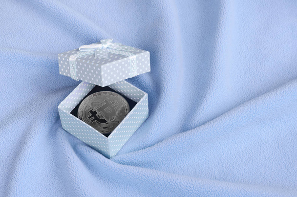 Stříbrný bitcoin leží v malé modré krabičky s malý luk na deku z měkké a nadýchané světle modrá fleece tkaniny s velkým počtem úlevu záhyby - Fotografie, Obrázek