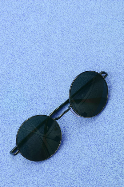Стильні чорні сонцезахисні окуляри з круглими окулярами лежать на ковдрі з м'якої та пухнастої світло-блакитної тканини флісу. Модна фонова картина в жіночих кольорах
 - Фото, зображення