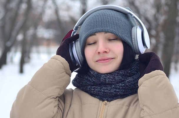 Красивая девушка носит теплые зимние наушники и шарф. Веселая брюнетка-модель гуляет на улице в наушниках в холодную погоду. Улыбающаяся счастливая модель позирует. Хорошая погода для отдыха
 - Фото, изображение