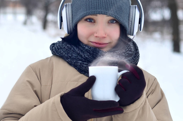 Bella forata bruna caucasica giovane donna sorridente mentre ascolta musica su grandi cuffie bianche musica e bere una tazza di caffè nel parco invernale o nella foresta
 - Foto, immagini