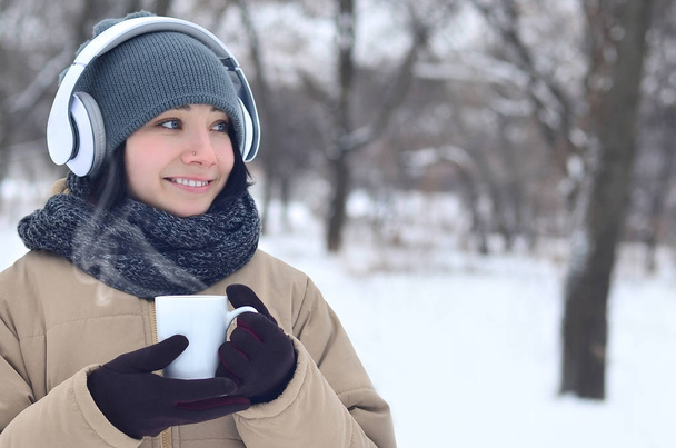 Portrait hivernal d'une jolie caucasienne percée de bois enneigé. Elle écoute de la musique sur de grands écouteurs blancs avec une tasse de café dans ses mains. Promenades en plein air
 - Photo, image