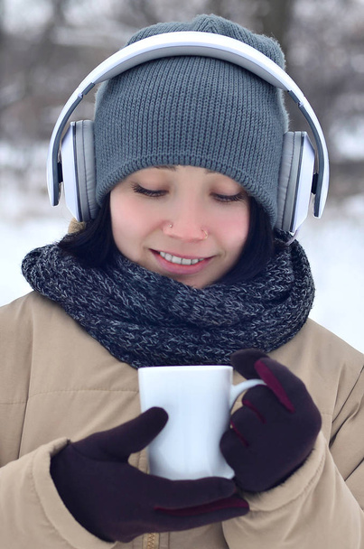 Beautiful pierced brunette caucasian young woman smiling while listening to music on large white headphones music and drink a cup of coffee in winter park or forest - Photo, Image