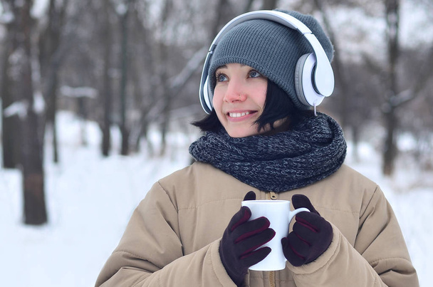 Winter portrait of pretty attractive caucasian pierced girl in snowy wood. She is listening to music on big white headphones with coffee cup in her hands. Walks in the open air - Photo, image
