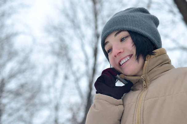 冬の森の美しさの少女。美しい若いブルネット白人ピアス女性おしゃれな茶色のコートと暖かい灰色の帽子でスマート フォンを使用して - 写真・画像