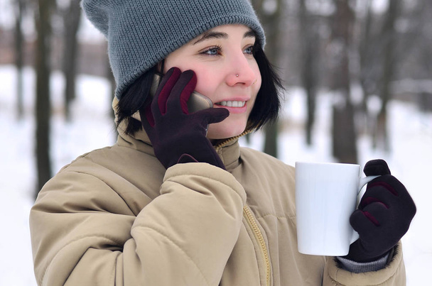 Porträt einer hübschen kaukasischen gepiercten Hipsterin im Winterpark. Sie spricht auf einem modernen Smartphone und trinkt Kaffee. Jugend-Straßenmode. Spaziergänge im Freien - Foto, Bild