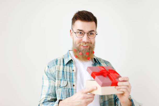 Fashionable bearded hipster guy in eyewear, opens wrapped present, recieves surprise from wife, being in good festive mood, isolated over white background. People, surprisment and presents concept - Foto, Bild