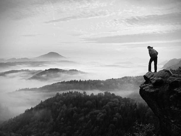 El hombre permanece en el pico rocoso durante el amanecer y vigila el paisaje brumoso
. - Foto, Imagen