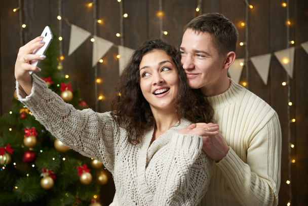 Щаслива пара приймає селфі і розважається в різдвяних прикрасах. Темний дерев'яний інтер'єр зі світлом. Романтичний вечір і концепція любові. Новорічне свято
. - Фото, зображення