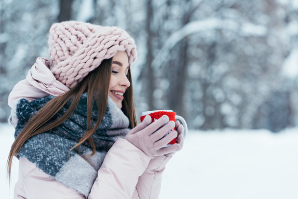 side view of beautiful young woman with cup of hot coffee in hands in snowy park - Photo, Image