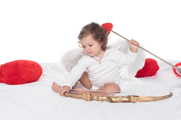 małego aniołka siedzącego na łóżku z łukiem i strzałami, na białym tle - Zdjęcie, obraz