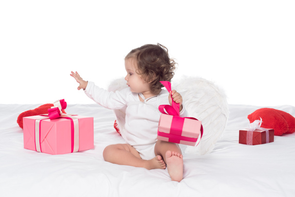 χαριτωμένο μωρό Άγγελος κάθεται στο κρεβάτι με δώρα, που απομονώνονται σε λευκό - Φωτογραφία, εικόνα