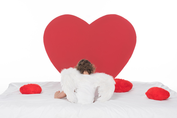 little cherub with wings lying on bed with hearts, isolated on white - 写真・画像