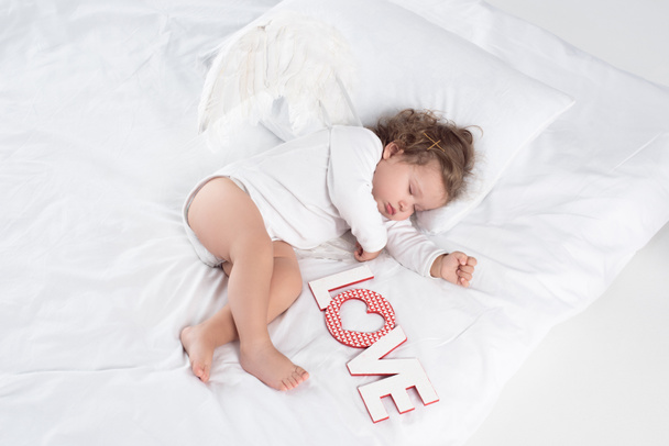 μικρό αγγελούδι με φτερά ξαπλωμένος στο κρεβάτι με το σύμβολο της αγάπης - Φωτογραφία, εικόνα