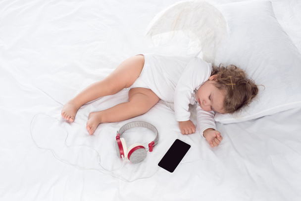 λίγο Άγγελος με φτερά ξαπλωμένος στο κρεβάτι με τα ακουστικά και smartphone - Φωτογραφία, εικόνα