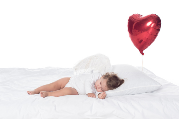 malý andílek s křídly spaní na posteli s balónky-srdce, izolované na bílém - Fotografie, Obrázek