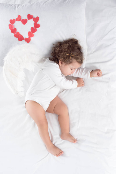 adorable cupid with wings sleeping on bed with red hearts - Foto, afbeelding