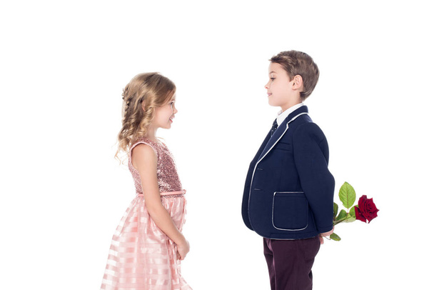 вид сбоку на модного мальчика в костюме, держащего розовый цветок и смотрящего на восхитительную маленькую девочку в розовом платье, изолированном на белом
  - Фото, изображение