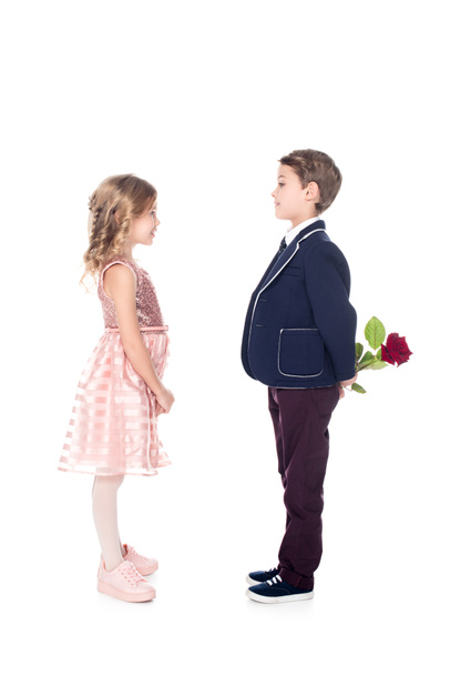 Seitenansicht des stilvollen Jungen im Anzug mit Rosenblume und Blick auf schöne kleine Mädchen in rosa Kleid isoliert auf weiß  - Foto, Bild