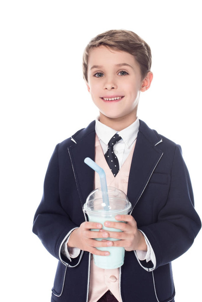 süßer kleiner Junge hält Milchshake im Plastikbecher und lächelt in die Kamera isoliert auf weiß - Foto, Bild