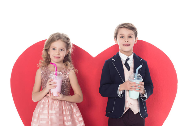 niedliche kleine Kinder mit Milchshakes in Plastikbechern und großem roten Herz hinter isoliert auf weiß - Foto, Bild