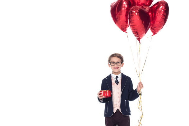 sevimli küçük çocuk takım elbise ve kırmızı kalp şeklinde balonlar ve üzerinde beyaz izole hediye kutusu tutarak gözlük - Fotoğraf, Görsel