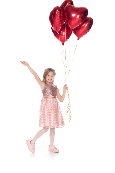 hermoso niño en vestido rosa sosteniendo globos en forma de corazón y sonriendo a la cámara aislada en blanco
 - Foto, Imagen